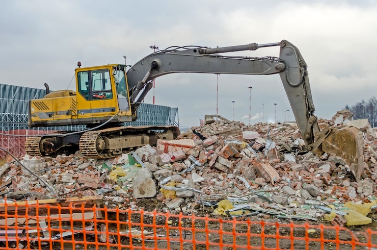 A large yellow-and-gray excavator sits atop a towering pile of broken bricks, concrete slabs, and construction debris under an overcast sky. Its long articulated arm extends forward, with the shovel resting near the base of the rubble. An orange safety fence runs across the foreground, partially blocking the bottom of the pile.