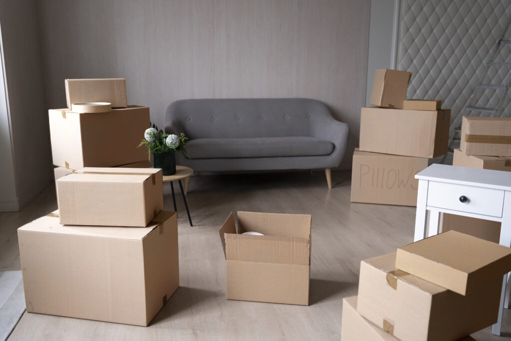 A living room in the process of being moved into, featuring a gray sofa surrounded by stacked cardboard moving boxes. Boxes are placed near a window on the left and stacked on the right, including one labeled "PILLOWS." A small round table with a potted plant sits between the sofa and boxes, and a white side table with a drawer stands on the right side of the room. The room has light wood flooring and light neutral walls.