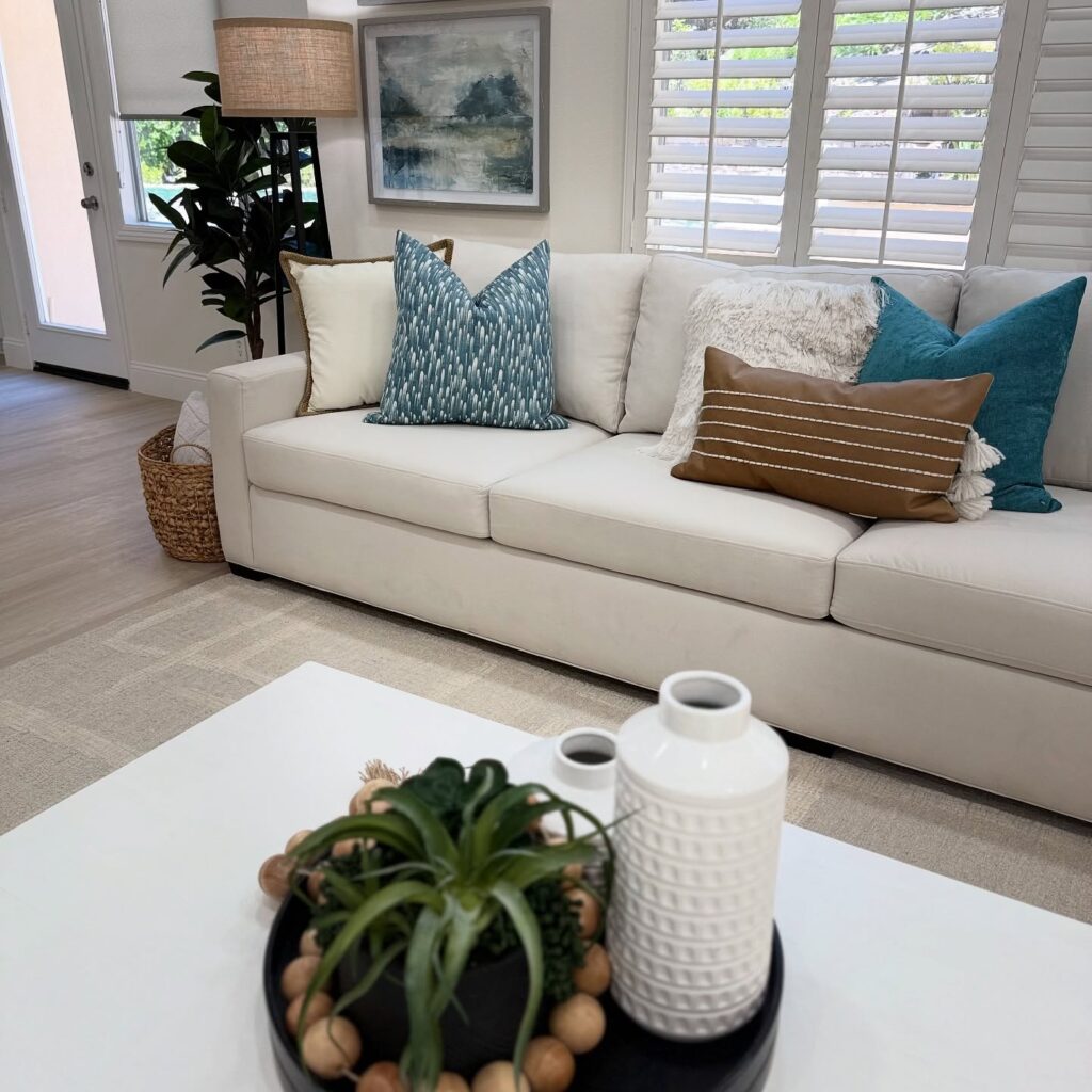 Bright living room with an off-white sectional sofa, blue and cream throw pillows, window with white shutters, tall potted plant, framed landscape art, and a coffee table with decor.