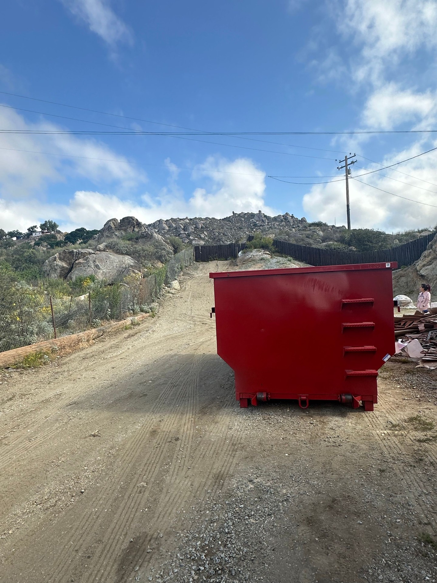 A large red dumpster sits on a dirt road, its side featuring horizontal metal bars like a ladder. The unpaved road curves uphill to the left behind it, bordered by a low chain-link fence and a rocky, brush-covered hillside with scattered vegetation.