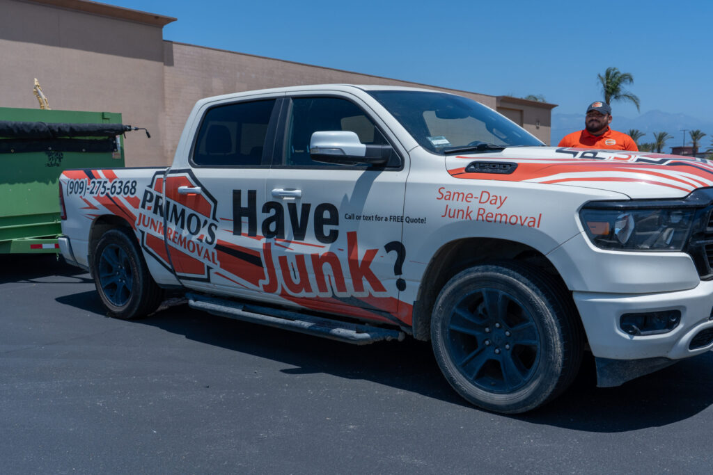 A white pickup truck branded with "Primo's Junk Removal" and red-and-black "Have Junk?" graphics, hitched to a green trailer, parked outside on a sunny day with palm trees, a building, and a man in an orange shirt and cap visible in the background under a clear blue sky.