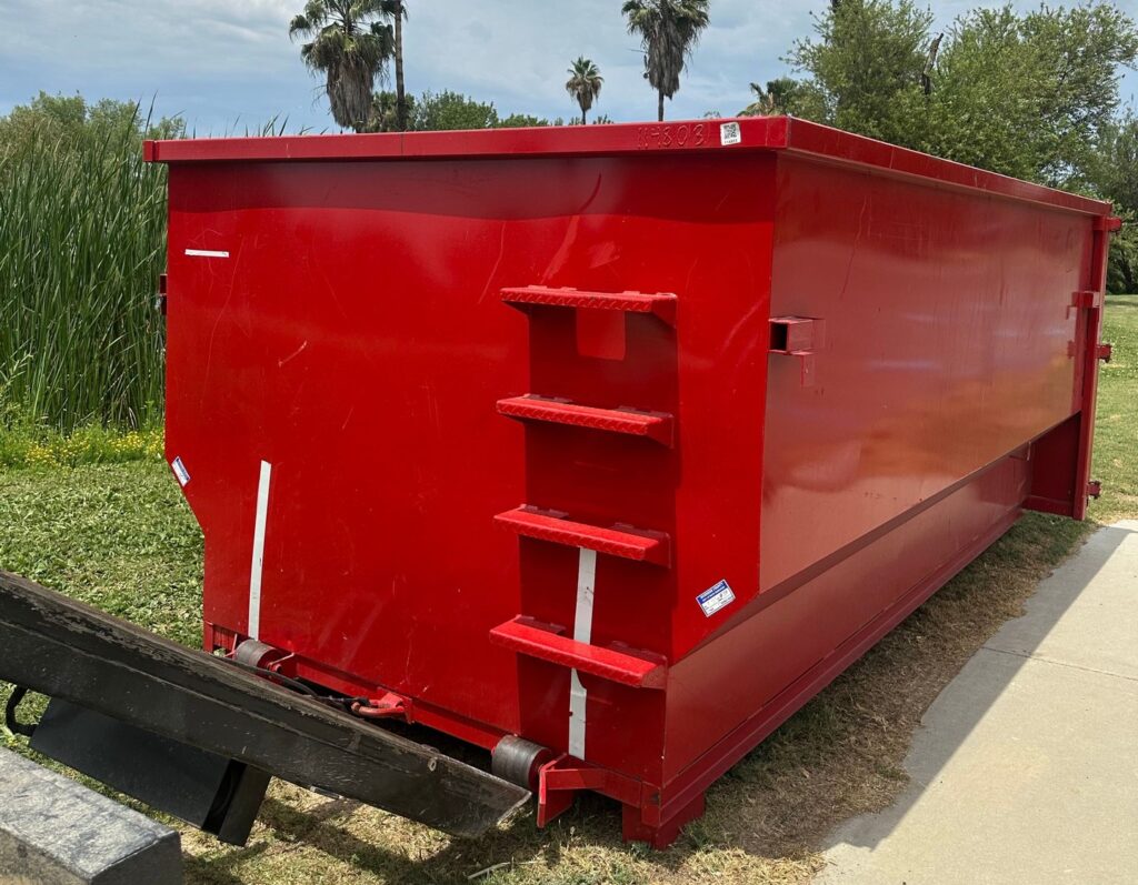 A large red dumpster sitting on concrete and grass, with three metal steps on its side. The background shows tall reeds, palm trees, and other greenery under a cloudy sky. A black ramp is partially visible in the foreground.