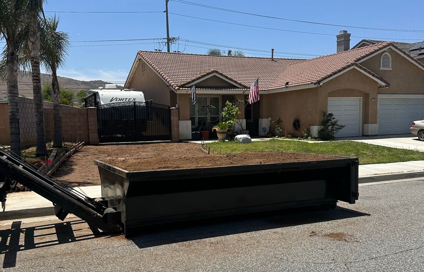 A dumpster filled with dirt sits on a residential street in front of a single-story stucco house with a red-tiled roof under a clear blue sky. A mechanical arm extends from the dumpster toward the left. An American flag hangs near the home's entrance. Behind a metal gate to the left, a white RV marked "VORTEX" is parked. Palm trees and greenery surround the property.