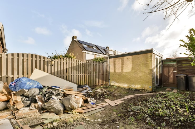 A backyard scene with a large pile of trash and debris on the left side, including blue and black garbage bags, cardboard boxes, and wooden planks. The right side of the yard is bare dirt with sparse green vegetation.
