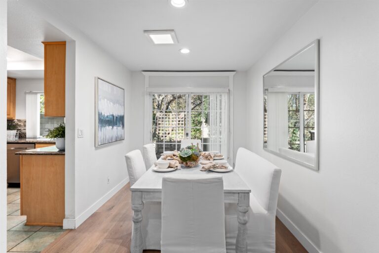 Bright dining room with white table set for a meal, succulent centerpiece, abstract blue painting on wall, mirror reflecting greenery, window with tree view, and kitchen beyond.