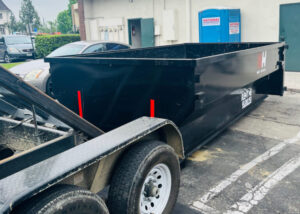 A large black roll-off dumpster being towed through an asphalt parking lot on a trailer. The dumpster is empty with its lid slightly open, featuring a red 'H' logo with 'WE HAUL' printed beneath it and 'RENT ME ROLLING' text along its side.
