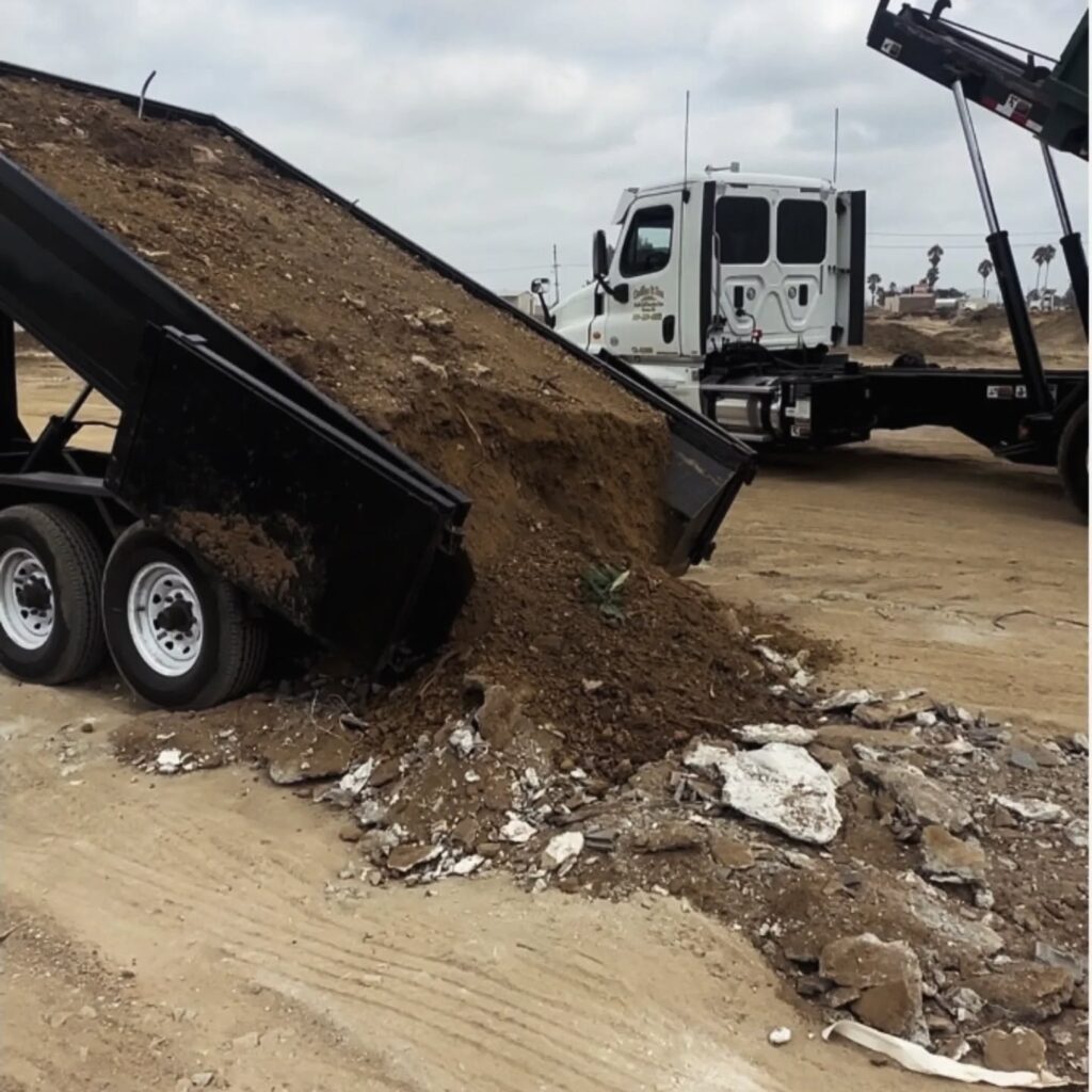 A dump truck with a partially raised bed is tipping a large load of brown dirt and construction debris onto sandy ground, where it mixes with chunks of white and gray concrete. The truck has a white cab and chassis, and the sky is overcast. In the background, a flatbed truck with a raised hook lift system is visible.