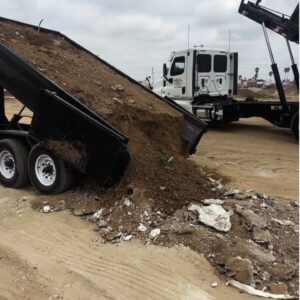 A dump truck with a partially raised bed is tipping a large load of brown dirt and construction debris onto sandy ground, where it mixes with chunks of white and gray concrete. The truck has a white cab and chassis, and the sky is overcast. In the background, a flatbed truck with a raised hook lift system is visible.