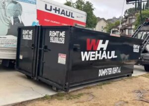 A large black dumpster with "WH WEHAUL DUMPSTER RENTAL" in white and red lettering sits in a driveway, with a white U-Haul truck visible behind it, houses and green trees under an overcast sky in the background.
