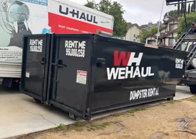 A large black dumpster with "WH WEHAUL DUMPSTER RENTAL" in white and red lettering sits in a driveway, with a white U-Haul truck visible behind it, houses and green trees under an overcast sky in the background.
