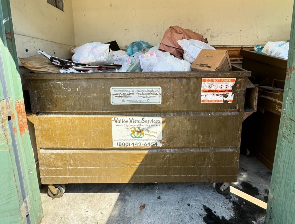 A large olive-green industrial dumpster overflowing with trash sits in a concrete waste enclosure. The dumpster is piled high with white plastic bags, cardboard boxes, and assorted debris.