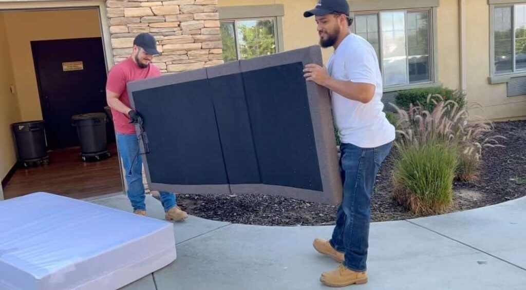 Two men in t-shirts and jeans carry a large, dark upholstered headboard outdoors, walking across the exterior of a stone-clad building toward a doorway. A white mattress wrapped in clear plastic lies in the foreground, and ornamental grass and windows appear in the background.
