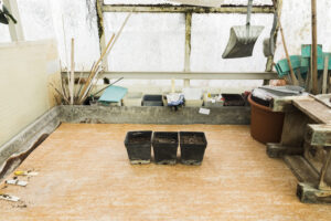 A weathered greenhouse interior with translucent panels, wooden framing, and sand floor. Three small black pots sit in the foreground, while a stone planter box along the back wall holds wooden stakes, a blue cloth, and gardening tools.