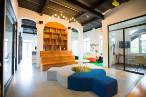 Modern open-plan office common area with a wooden shelf-lined archway, circular modular seating in grey, blue, and yellow, and colorful bean bags visible through glass partitions.