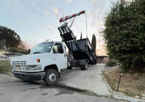 A white GMC dump truck with a raised black dumpster angled upward on a red-and-white striped hydraulic lift sits on a paved driveway, facing left.