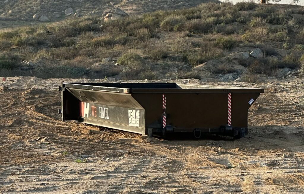 A dark brown "WE HAUL" dumpster with "RENT ME" text and white-and-red striped safety markers sits on dry dirt, backed by a sparse brush-covered hillside.