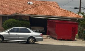 A silver sedan parked on the street beside a house with an open garage, where a large red dumpster sits in the driveway and extends into the garage opening.