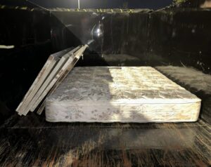 An old, stained floral-patterned mattress sits inside a dark dumpster, beside a stack of four light rectangular panels with brown marks, against scratched wood and black metal walls under bright overhead light.