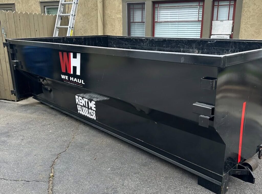 A large empty black dumpster with white "W.H. WE HAUL" logo and red stripe sits on an asphalt driveway, advertising rental phone number, with a building and wood fence behind.