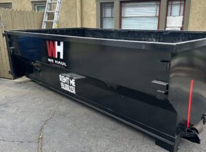 A large empty black dumpster with white "W.H. WE HAUL" logo and red stripe sits on an asphalt driveway, advertising rental phone number, with a building and wood fence behind.